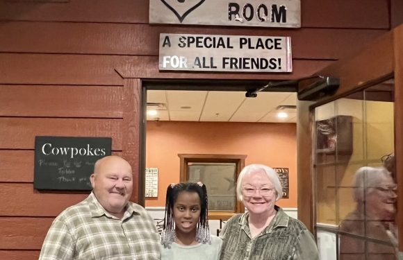 Three people stand together inside a room with wooden paneling. They are smiling and posing in front of a doorway, above which is a sign that reads "Abbie's Room: A special place for all friends!".