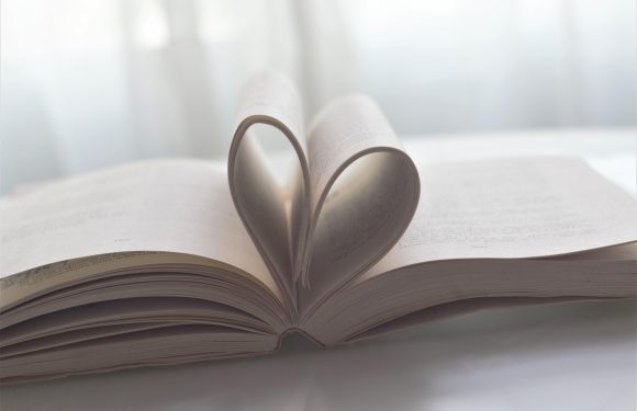 An open book with pages curled to form a heart shape, sitting on a white surface with a soft, bright background.