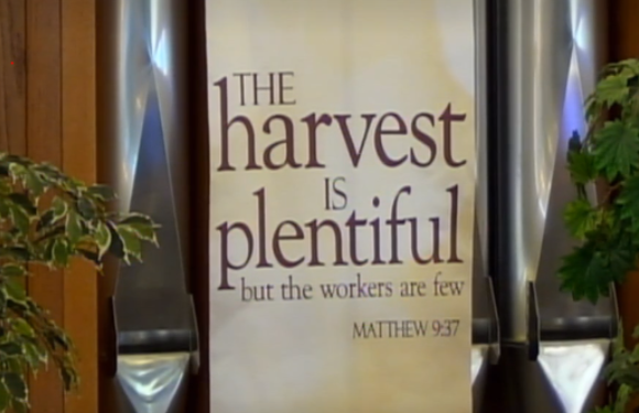 A religious banner reads "The harvest is plentiful but the workers are few" with a Bible reference, hanging between wooden panels and flanked by plants.
