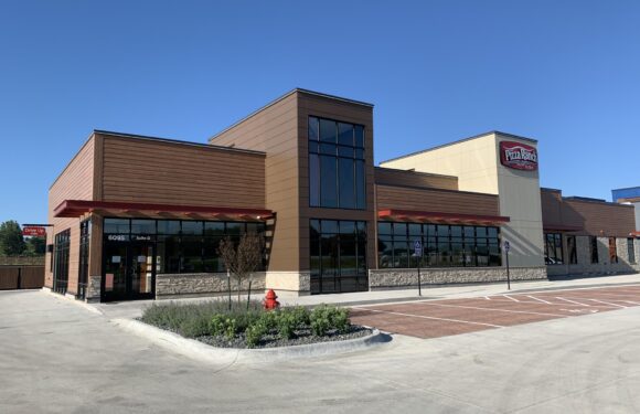 A modern, single-story building with large windows, brown and beige exterior, red awnings, and a Pizza Ranch sign, with a clear blue sky overhead.