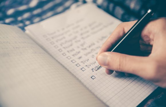 A person's hand holding a black pen, checking off items on a handwritten to-do list in a grid notebook with a blurred background.