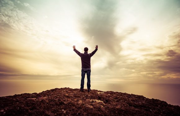 A person stands on rocky terrain at sunset, arms raised toward a cloudy sky, with a calm ocean visible in the background.
