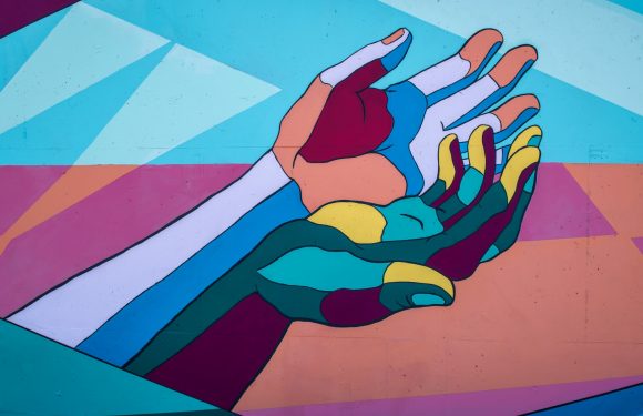 A colorful mural of two stylized hands, one with a light skin tone and the other with a rainbow pattern, reaching towards each other against an abstract background.