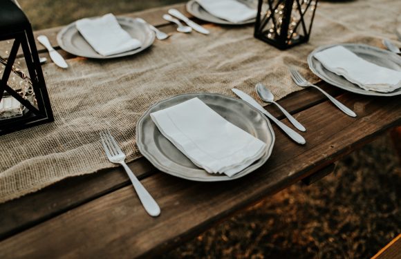 A rustic outdoor table set with white plates, silverware, and folded napkins, decorated with a burlap table runner and black lanterns with candles.