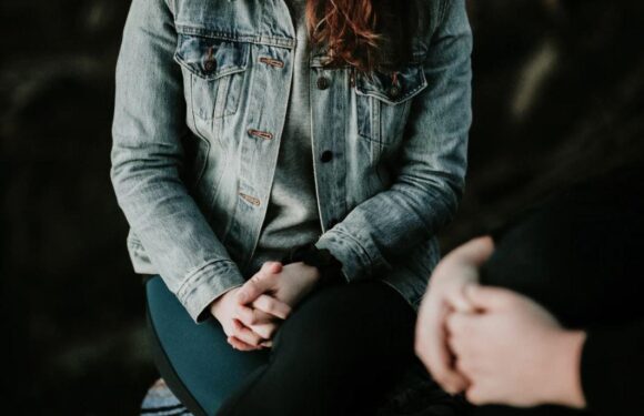 A person wearing a denim jacket and gray shirt sits with hands clasped on their lap, engaging in conversation with another person whose hands are visible.