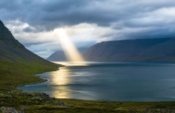 A serene landscape with mountains, a lake, and a sunbeam breaking through dark clouds illuminating part of the water and shoreline.