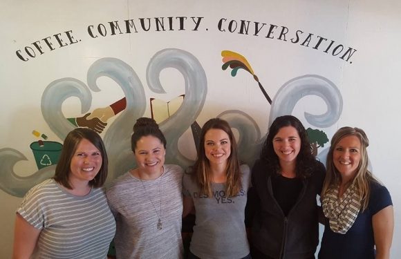 Five women standing together in front of a wall with a mural that includes the words "Coffee. Community. Conversation." and various illustrations.