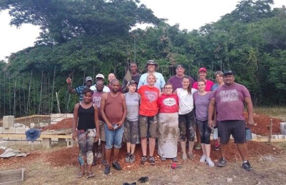 Group of diverse people standing outdoors on a construction site with trees in the background. They are smiling and some are making gestures.