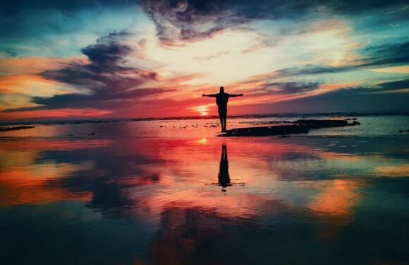 A person with arms outstretched stands on a rocky shoreline at sunset, with a colorful sky reflected in the calm water below.