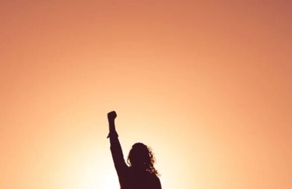 Silhouette of a person raising a fist in front of a bright, orange sunset sky. The individual has curly hair and is wearing a long-sleeve shirt or jacket.