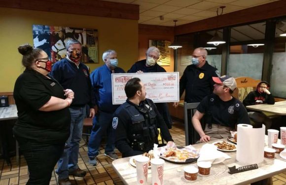 Group of people, including police officers and civilians, gathered inside a restaurant holding a large check. Some are wearing masks, and food is on the table.