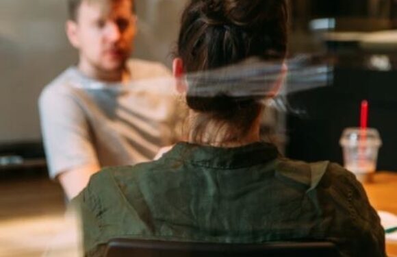 A woman with dark hair tied up in a bun, wearing a green jacket, sits at a table facing a man with blond hair in a casual gray t-shirt, with a blurred background.