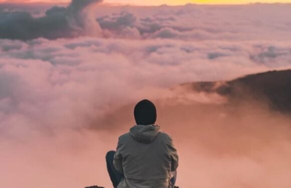 Person sitting on a rock at a mountaintop, overlooking a sea of clouds during sunset, with pastel pink, orange, and yellow sky.