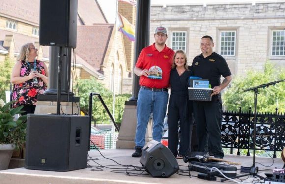Three people standing on a stage outdoors, smiling and posing for a photo, with a woman holding a microphone and two men holding a basket and an item.