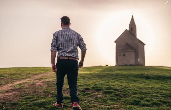 A man walking on a grassy hill toward a small church at sunset, wearing a checkered shirt, dark pants, and red shoes. The church has a steeple and stone walls.