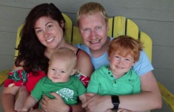 A smiling family of four sits on a yellow bench against a gray wall, with two young children and two adults, the woman has dark curly hair and the man has short blond hair.