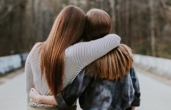 Two girls with long hair embrace each other with their arms around each other's shoulders on a pathway surrounded by trees.
