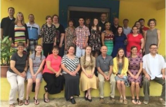 Group of 20 adults standing and sitting in front of a yellow and blue building, some wearing casual clothes and one person in a uniform with a Pizza Ranch logo.