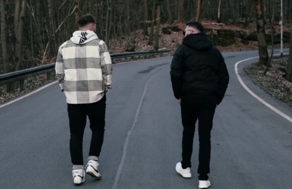 Two individuals walk down a winding, empty road surrounded by trees, wearing casual jackets and sneakers, with one in a plaid-patterned hoodie.