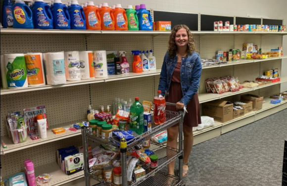 A woman with curly hair in a denim jacket and brown dress shops for groceries in a store aisle with shelves of cleaning products, paper towels, and snacks.