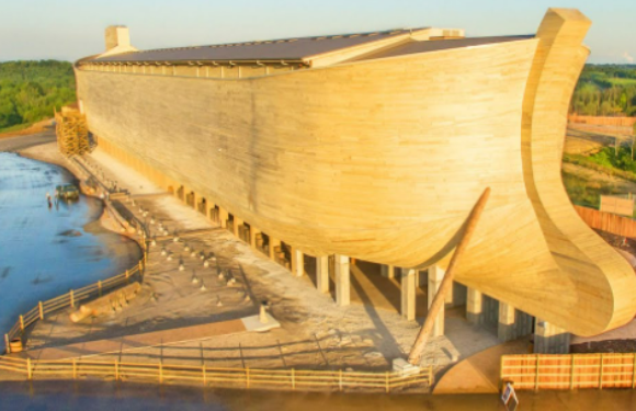 A large boat-shaped building with a wooden exterior, situated near a river, with green hills and clear sky in the background.