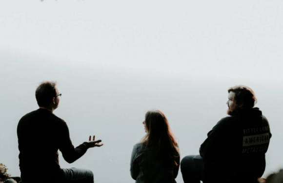 Three people are sitting outdoors against a light sky; two men and a woman are engaged in conversation, with one man gesturing with his hand.