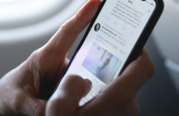 A person holding a smartphone, browsing Twitter, seated by an airplane window with natural light illuminating their hand and device.