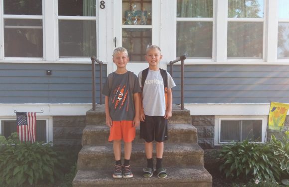 Two young boys with backpacks stand on the front steps of a house, smiling. They are dressed casually, with one wearing a Pizza Ranch T-shirt. The house has blue siding and large windows, with American and fall-themed flags hanging nearby.