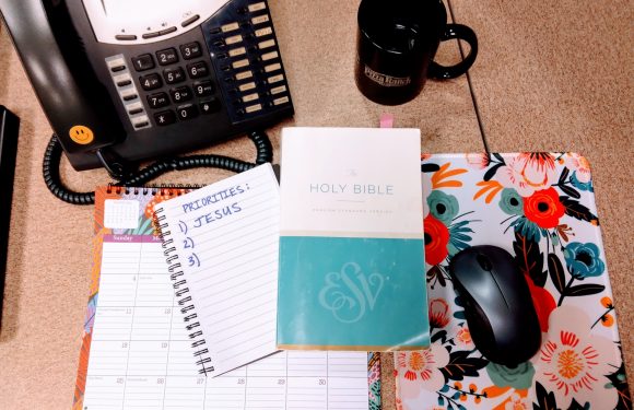 A desk with a black office phone, a black coffee mug labeled "Pizza Ranch," a floral desk mat, a computer mouse, a Holy Bible, a spiral notebook with "Priorities: 1. Jesus" written, and a floral weekly planner.
