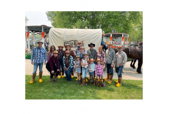 Group of people dressed in western attire with hats, boots, and jeans, standing in front of a wagon with a "Pizza Ranch" sign and a horse in the background.