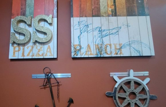 Two wall art pieces with a nautical theme hang on an orange wall. The left features large, glittery "SS" letters and "PIZZA" below, the right shows a blue sketch of a ship.