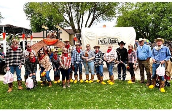 Group of people dressed in Western attire, holding ropes and wearing cowboy hats, standing in front of a decorated float with a "Pizza Ranch" sign, on a grassy area.
