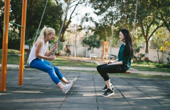 Two women are sitting on swings in a park, smiling and facing each other while enjoying a sunny day surrounded by trees.