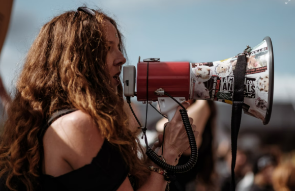 A woman with long, curly hair and a sleeveless top speaks into a megaphone decorated with various stickers during an outdoor event.