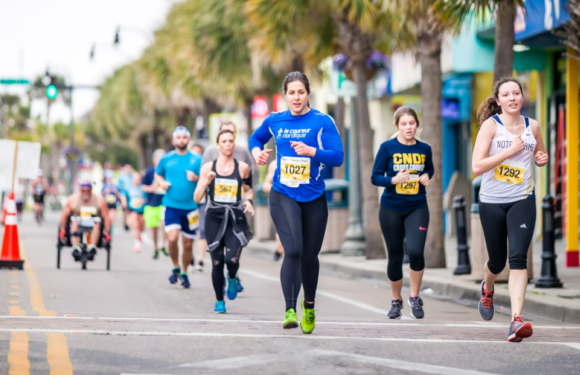 Group of runners participating in a race on a city street lined with palm trees, with some runners wearing race bibs and athletic clothing.