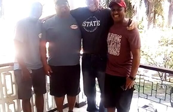Four people standing on a balcony, smiling, with palm trees in the background; the person on the left is wearing a light gray shirt and dark shorts.
