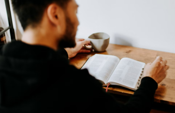 A person with short dark hair and a beard, wearing a black hoodie, reads a large open book at a wooden table, holding a beige bowl in one hand.