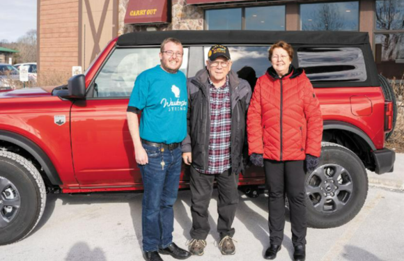 Three people stand in front of a red vehicle with a black roof, outdoors, wearing casual jackets, with a building and trees in the background.