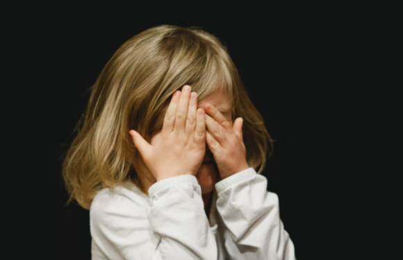 A young girl with blonde hair wearing a white long-sleeve shirt covers her face with both hands against a black background.