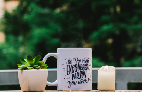 A white mug with the quote "be the most encouraging person you know" sits between a small potted succulent and a lit candle on a wooden surface, with a blurred green background.