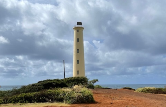 A tall, cream-colored lighthouse on a grassy and sandy coastal area under a blue sky with scattered clouds.