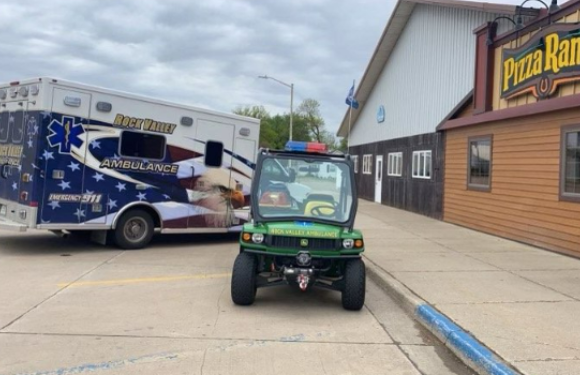 A green utility vehicle parked outside a Pizza Ranch, with an ambulance nearby on a mostly cloudy day. The building has a wooden exterior and signage.