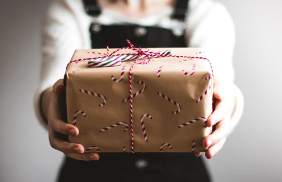Person holding a gift wrapped in brown paper with candy cane pattern, tied with red and white string, with a small black and white striped tag on top.
