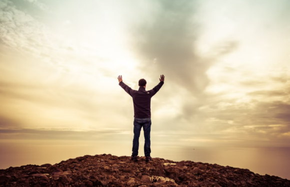 A person stands on rocky ground with arms raised, facing a cloudy sky during sunset or sunrise, creating a peaceful and triumphant scene.