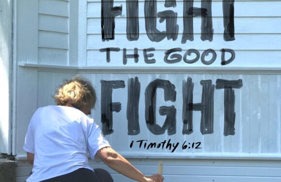 A person with curly hair in a white shirt and black pants is painting a sign on a white building, reading "FIGHT THE GOOD FIGHT" with a Bible verse reference at the bottom.