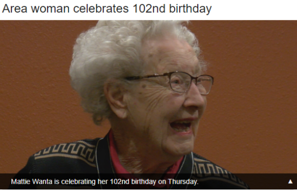 An elderly woman with white hair and glasses smiling, wearing a dark jacket with gold accents, standing in front of an orange wall.