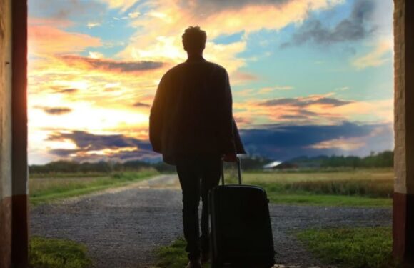 A person walking away with a rolling suitcase at sunset, seen through an arched doorway, with open fields and a dramatic sky in the background.