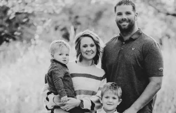 A family of four outdoors, with a woman holding a young child, standing next to a man and an older child, in a natural park setting with trees in the background.