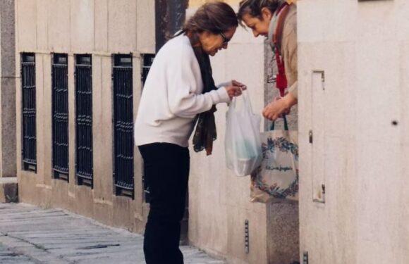 Two women exchange items on a sidewalk; one woman is in a white jacket, and the other is wearing an apron with floral patterns and holding a bag.
