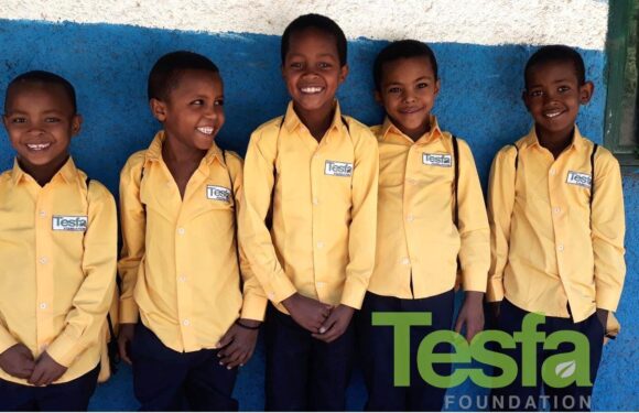 Six children in yellow shirts with "Tesfa Foundation" patches stand in a row against a blue and white wall, smiling and looking happy.